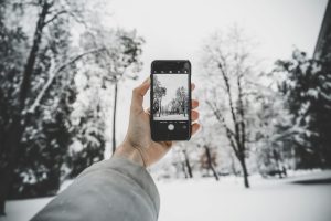 A hand holds a smartphone capturing a winter scene with snow-covered trees, showcasing the beauty of nature.